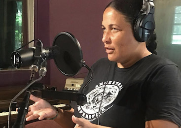 Woman wearing headphones and an Ancestral Imperative shirt speaks into a mic in a recording studio